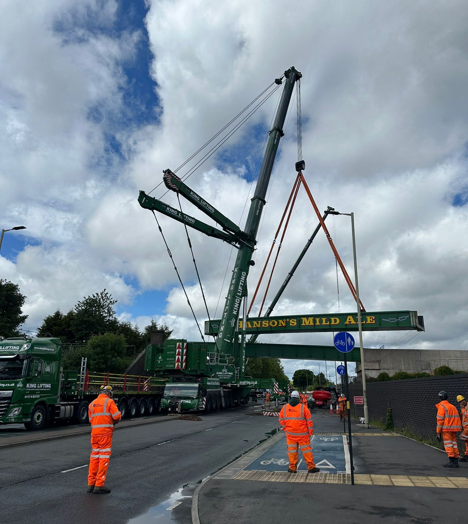 King Lifting crane team installing a Hanson's Mild Ale sign bridge with large green crane on urban street